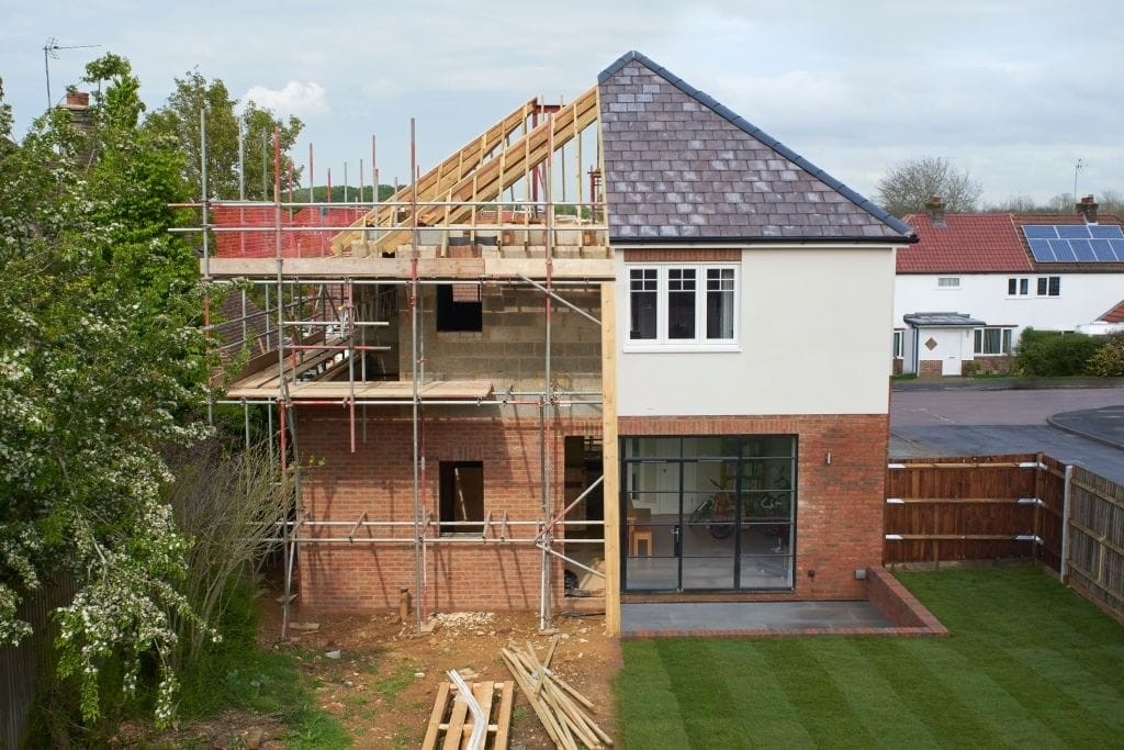 Construction site of a two-story house extension with scaffolding, showing a completed modern section with large glass doors and an unfinished brick structure, illustrating phases in residential development.