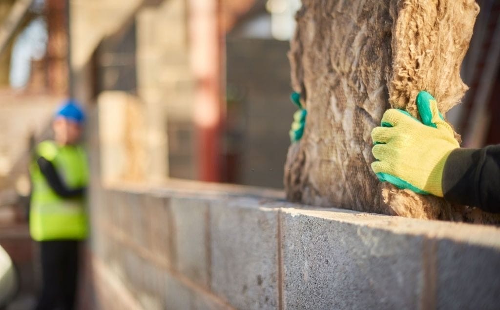 Close-up of a worker's hands with safety gloves carefully installing insulation material between concrete blocks on a construction site, with a supervisor in the background overseeing the work.